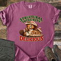 Structural Integrity: Delicious Comfort Colors T-shirt in Berry, laid flat. A cartoon beaver wearing a gingerbread roof as a hat nibbles a cookie beside a gingerbread log cabin on a tree stump, between the words Structural Integrity: Delicious. Olive pants and brown boots lie on a wood floor.