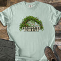 Freshwater Forest Comfort Colors T-shirt in Bay, laid flat. A driftwood arch draped in green moss shelters three small fish and bubbles above the words Freshwater Forest. The shirt lies on weathered wood boards beside folded olive pants and brown hiking boots.