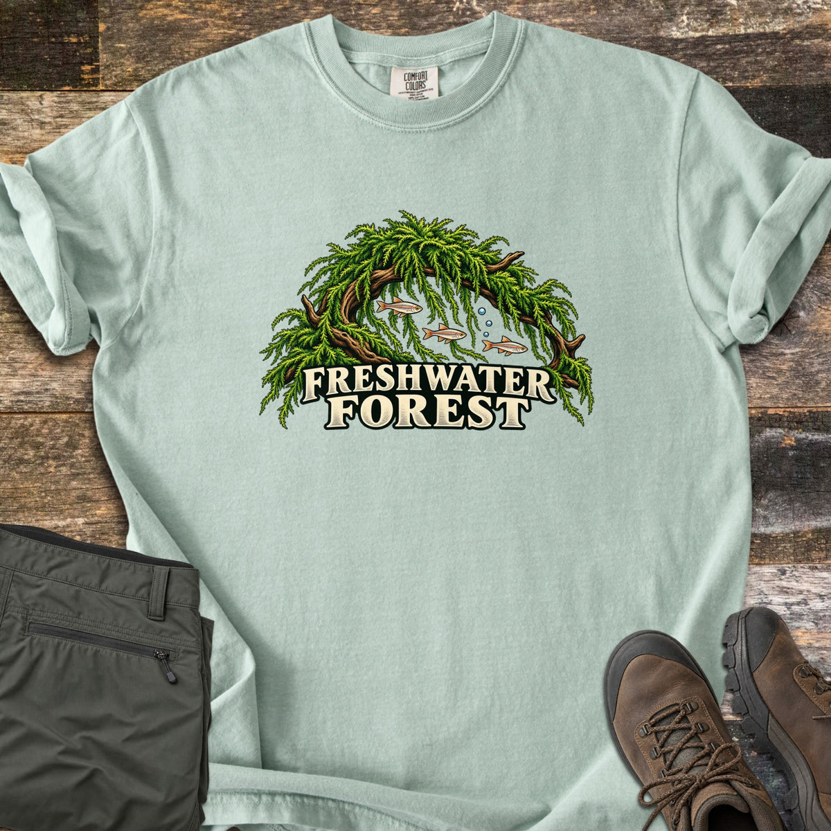 Freshwater Forest Comfort Colors T-shirt in Bay, laid flat. A driftwood arch draped in green moss shelters three small fish and bubbles above the words Freshwater Forest. The shirt lies on weathered wood boards beside folded olive pants and brown hiking boots.