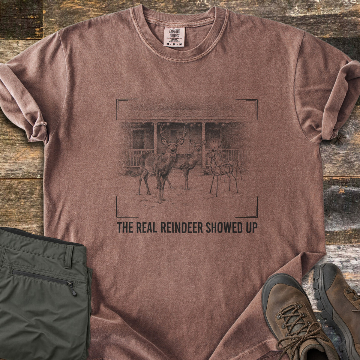 The Real Reindeer Showed Up Comfort Colors T-shirt in Espresso, laid flat. A sketch in camera viewfinder corners shows a buck and doe in snow by a wire reindeer decoration at a porch, above the words The Real Reindeer Showed Up. Olive pants and brown boots lie on weathered wood.
