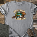 Watershed Engineer Comfort Colors T-shirt in Grey, laid flat. A beaver whose body frames a forest scene with mountains, a lake, and a waterfall flowing over a log dam, between the words Watershed Engineer. Olive pants and brown hiking boots lie on a wood surface.
