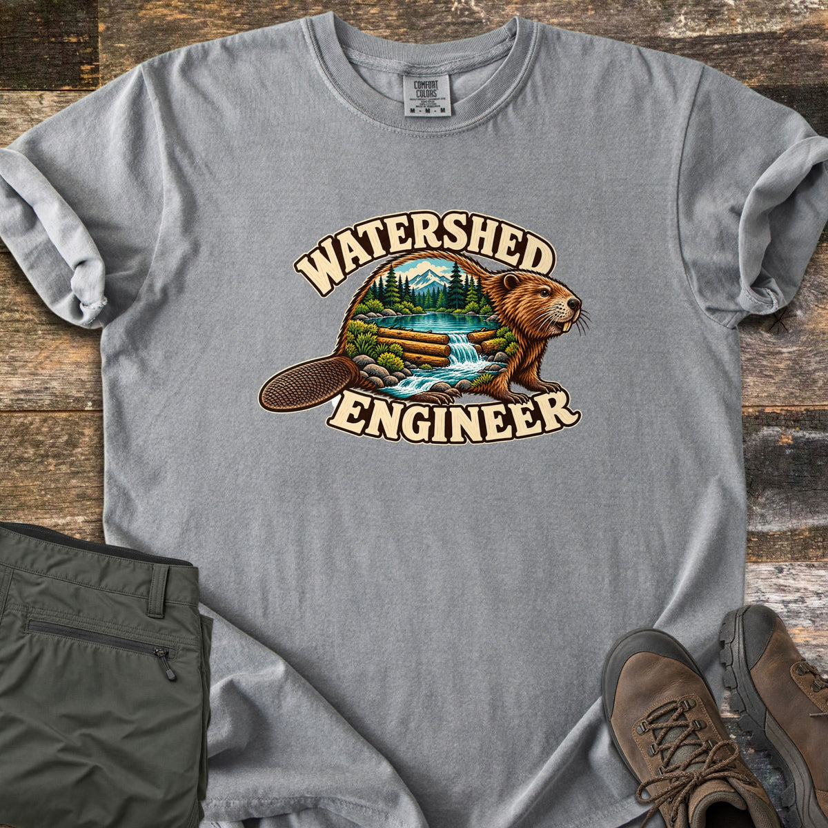 Watershed Engineer Comfort Colors T-shirt in Grey, laid flat. A beaver whose body frames a forest scene with mountains, a lake, and a waterfall flowing over a log dam, between the words Watershed Engineer. Olive pants and brown hiking boots lie on a wood surface.