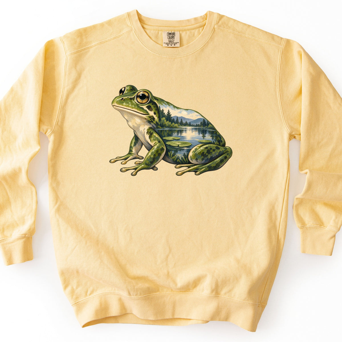 Lily Pond Frog Comfort Colors crewneck sweatshirt in Butter, laid flat. A green spotted frog sits in profile, its body filled with a scene of a calm lake, pine trees, distant mountains, and lily pads. No words are printed. The sweatshirt lies on a plain white background.