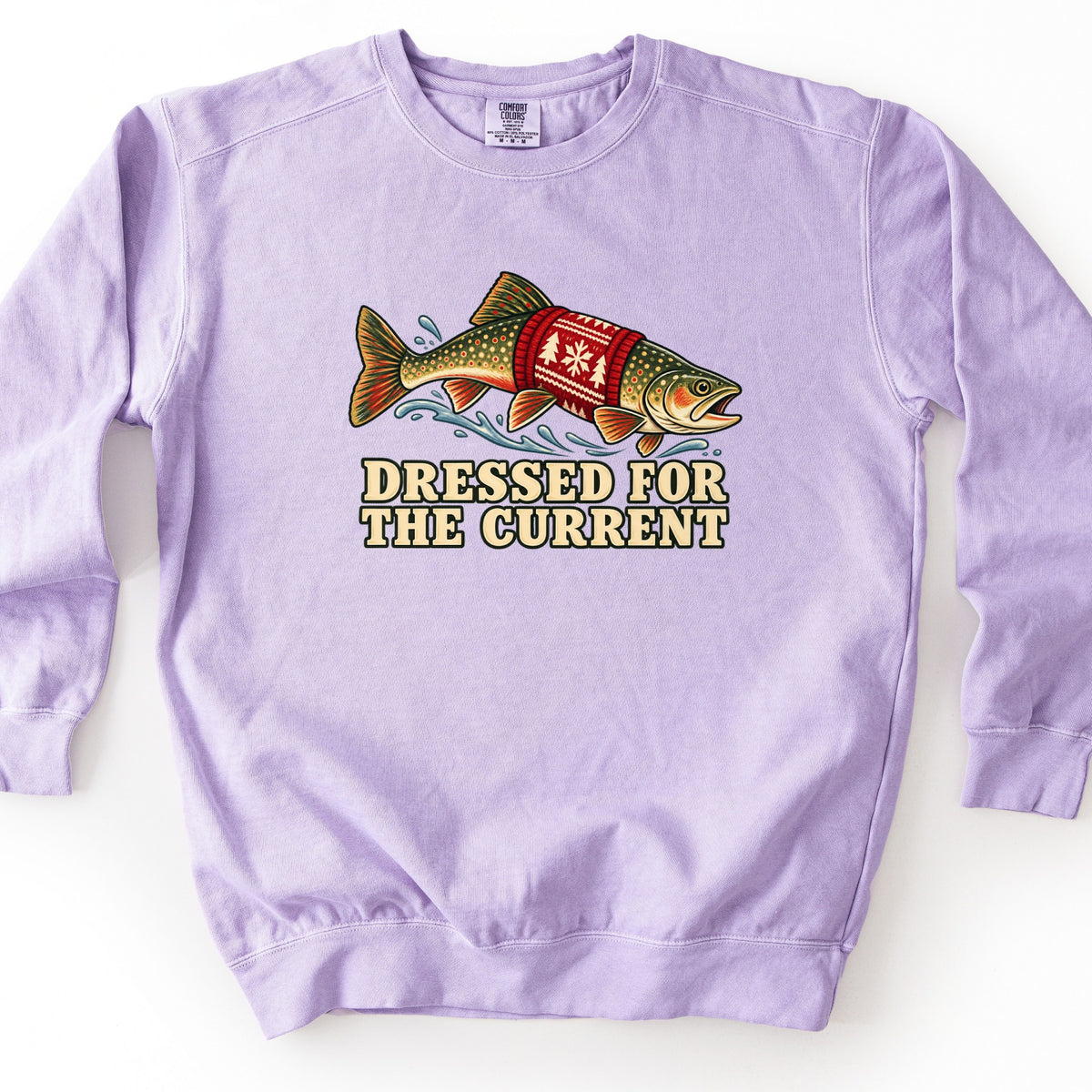 Dressed for the Current Comfort Colors crewneck sweatshirt in Orchid, laid flat. A spotted trout wearing a red knit holiday sweater with a tree and snowflake pattern leaps through splashing water above the words Dressed for the Current. Laid on a white background.