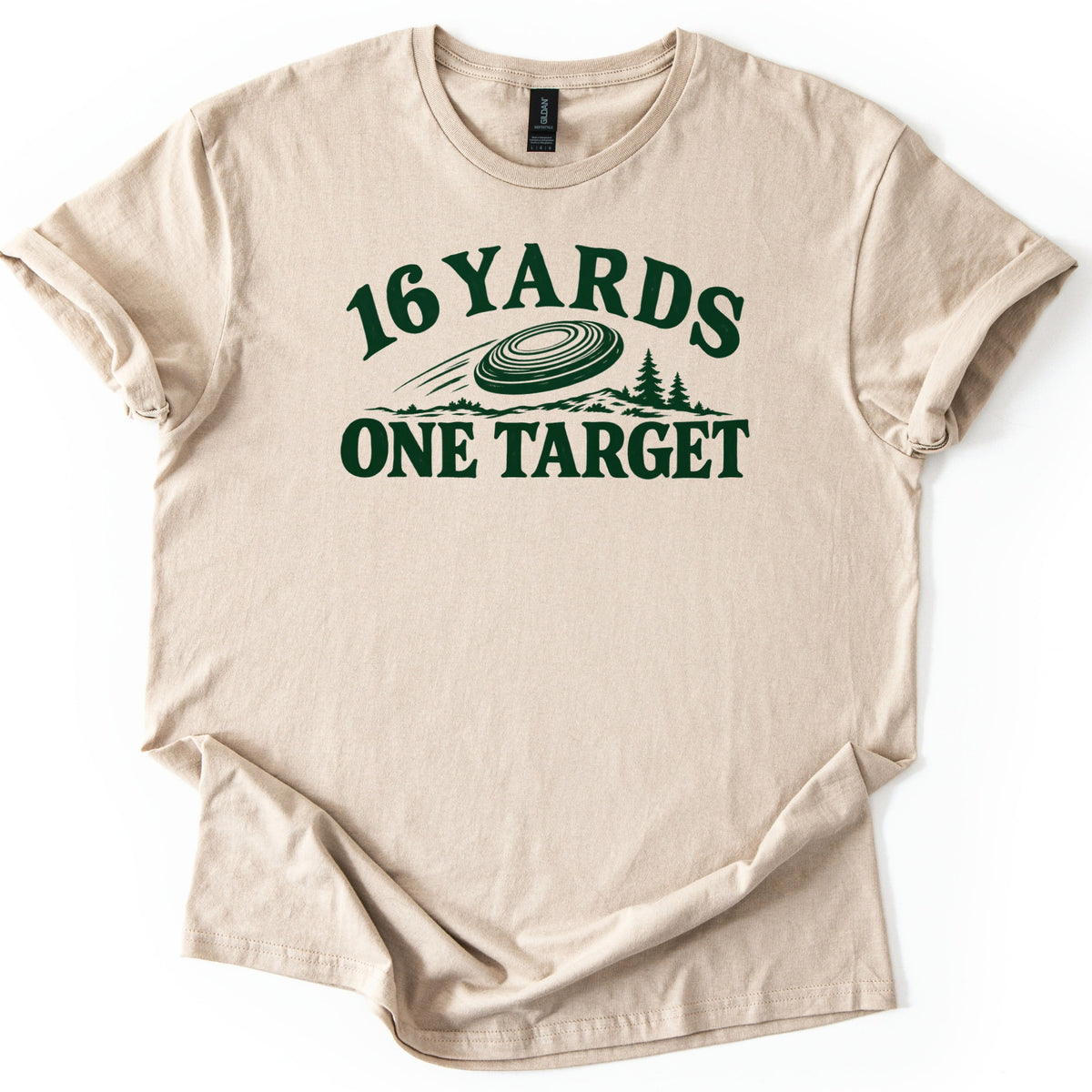 16 Yards One Target Comfort Colors T-shirt in Sand, laid flat. Dark green print of a flying clay target with motion lines over hills and pine trees, with 16 YARDS arched above and ONE TARGET below. The shirt has cuffed sleeves on a white background.