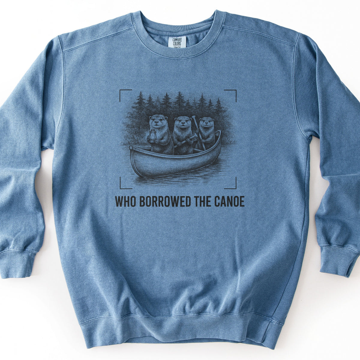 Who Borrowed The Canoe Comfort Colors crewneck sweatshirt in Blue Jean, laid flat. A black sketch of three otters in a canoe on a lake, one holding a lantern and two holding paddles, with pine trees behind and corner brackets around it. Below it reads WHO BORROWED THE CANOE. On white.