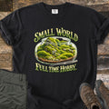 Small World Full Time Hobby Comfort Colors T-shirt in Black, laid flat. A glass jar terrarium filled with green moss, rocks, and pebbles, with metal tweezers beside it, between the words Small World and Full Time Hobby. On wood planks beside olive pants and brown hiking boots.