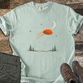 Moonrise Clay Comfort Colors T-shirt in Bay, laid flat. An orange clay target streaks past a crescent moon and small stars above pine trees and rolling hills, with no words. Folded olive cargo pants and brown hiking boots lie beside it on weathered wood boards.