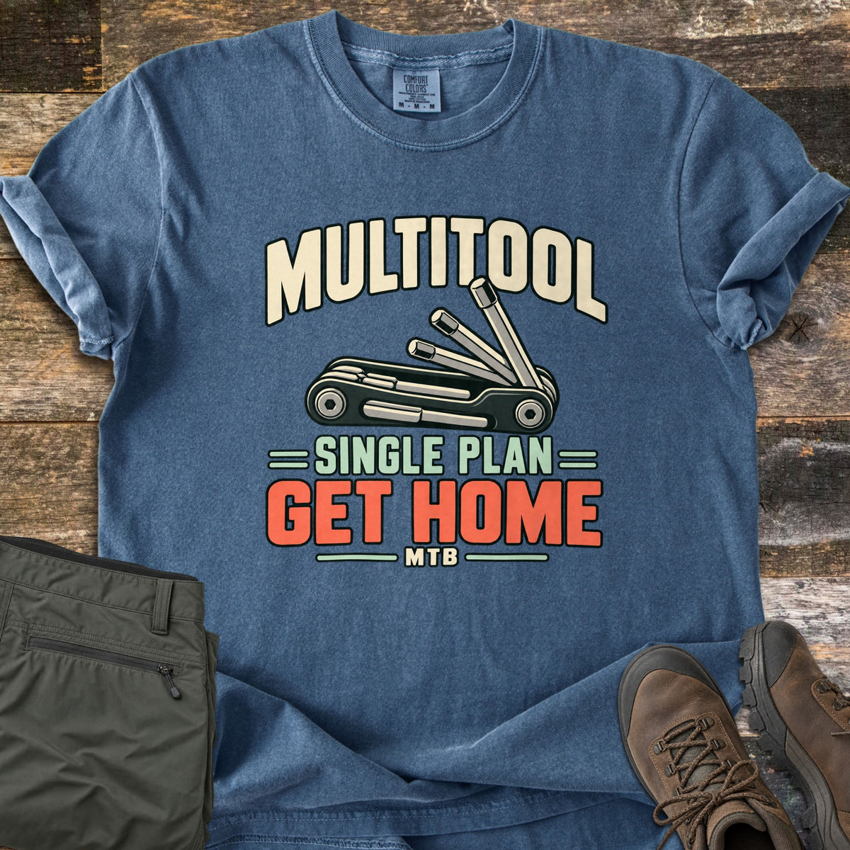 Multitool Single Plan Comfort Colors T-shirt in Denim, laid flat. An illustrated folding bike multitool with fanned-out hex keys sits under the word Multitool, above the words Single Plan Get Home MTB. Olive hiking pants and brown hiking boots lie beside it on weathered wood.