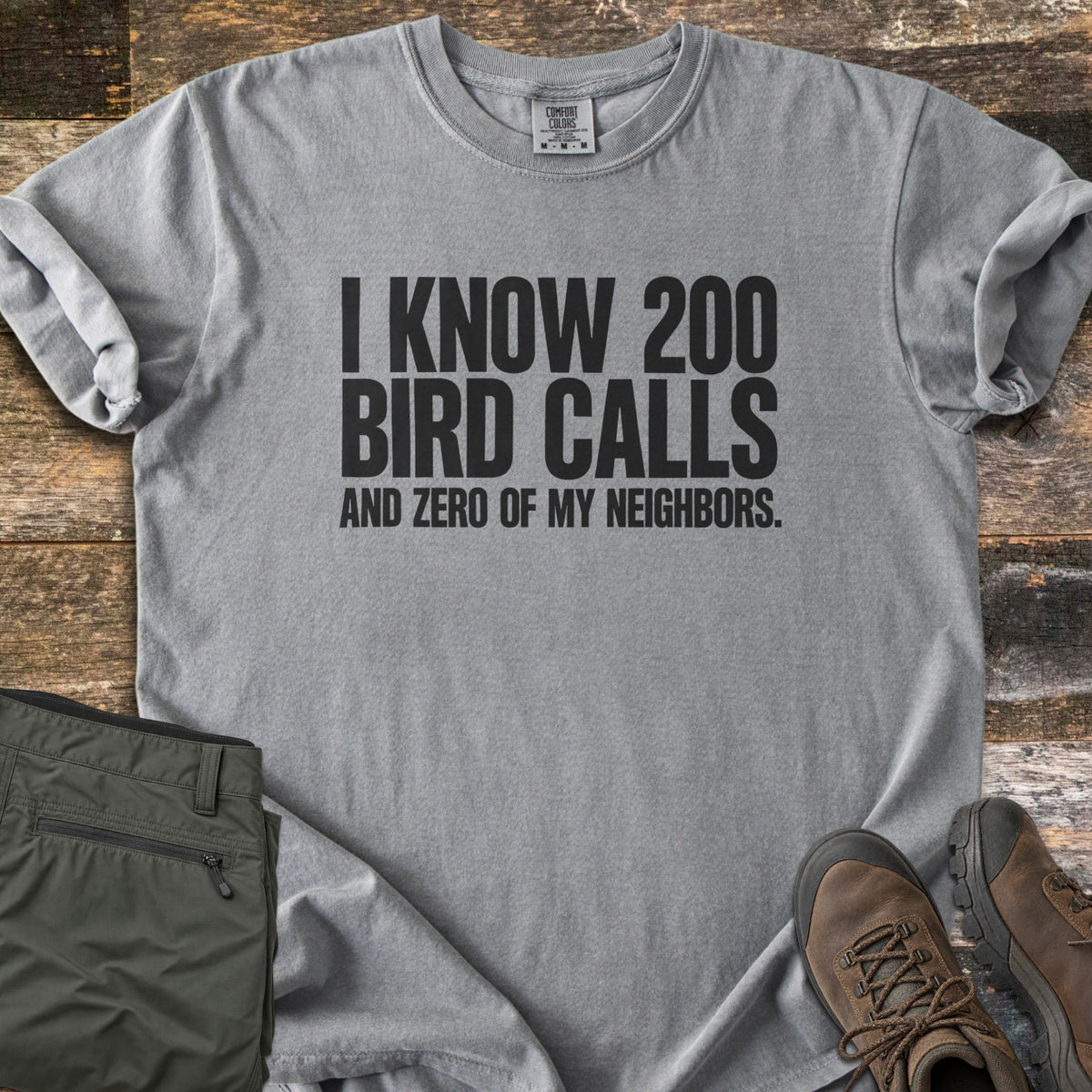 I Know 200 Bird Calls and Zero of My Neighbors Comfort Colors T-shirt in Grey, laid flat. Bold black capital letters read I KNOW 200 BIRD CALLS AND ZERO OF MY NEIGHBORS. The shirt has rolled sleeves and lies on weathered wood boards beside folded olive hiking pants and brown hiking boots.