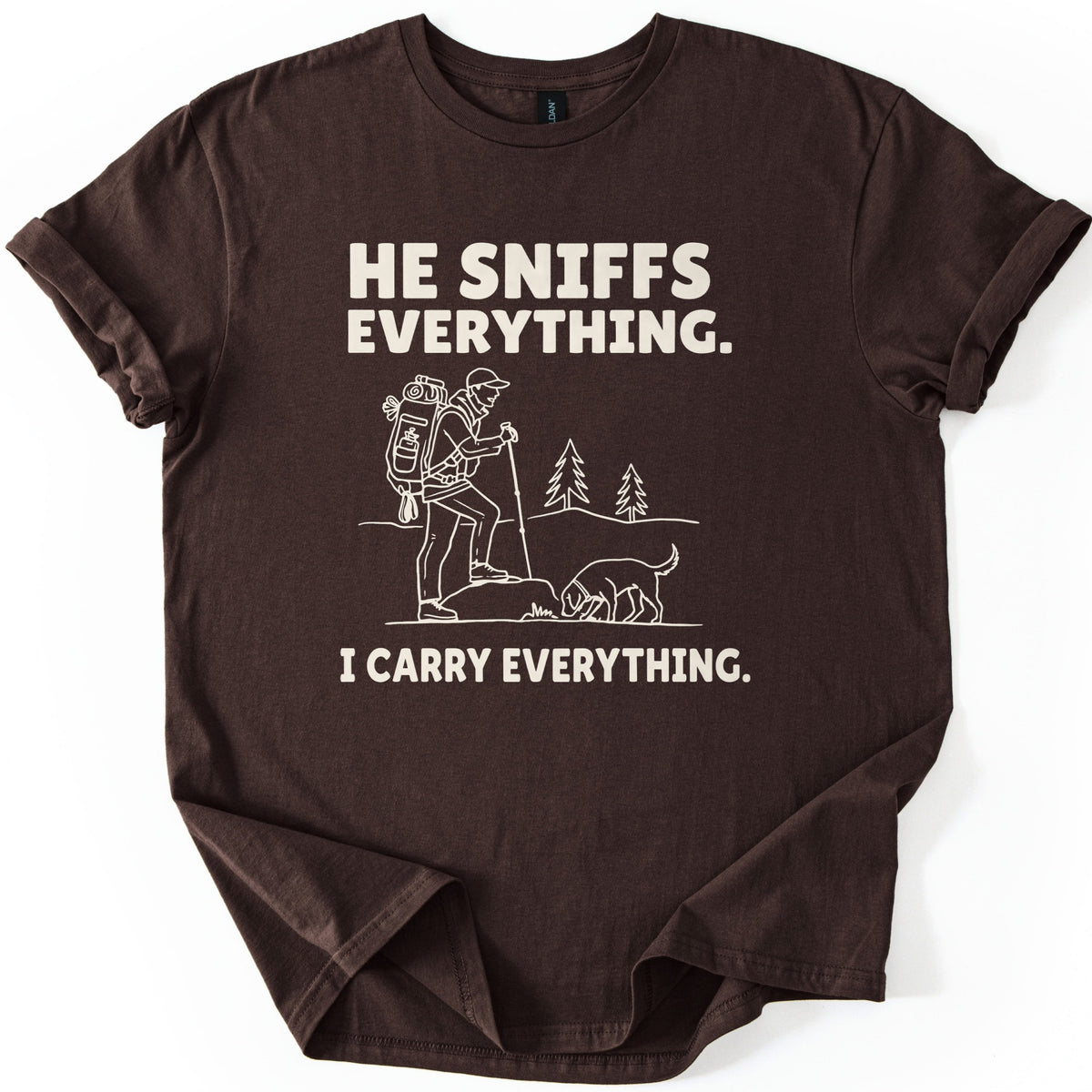 Trail Dog Pack Mule Comfort Colors T-shirt in Dark Chocolate, laid flat. Cream line art of a hiker with a loaded backpack and trekking pole stepping onto a rock as a dog sniffs the ground, with pine trees behind, between the words He sniffs everything. and I carry everything. Sleeves rolled, on white.