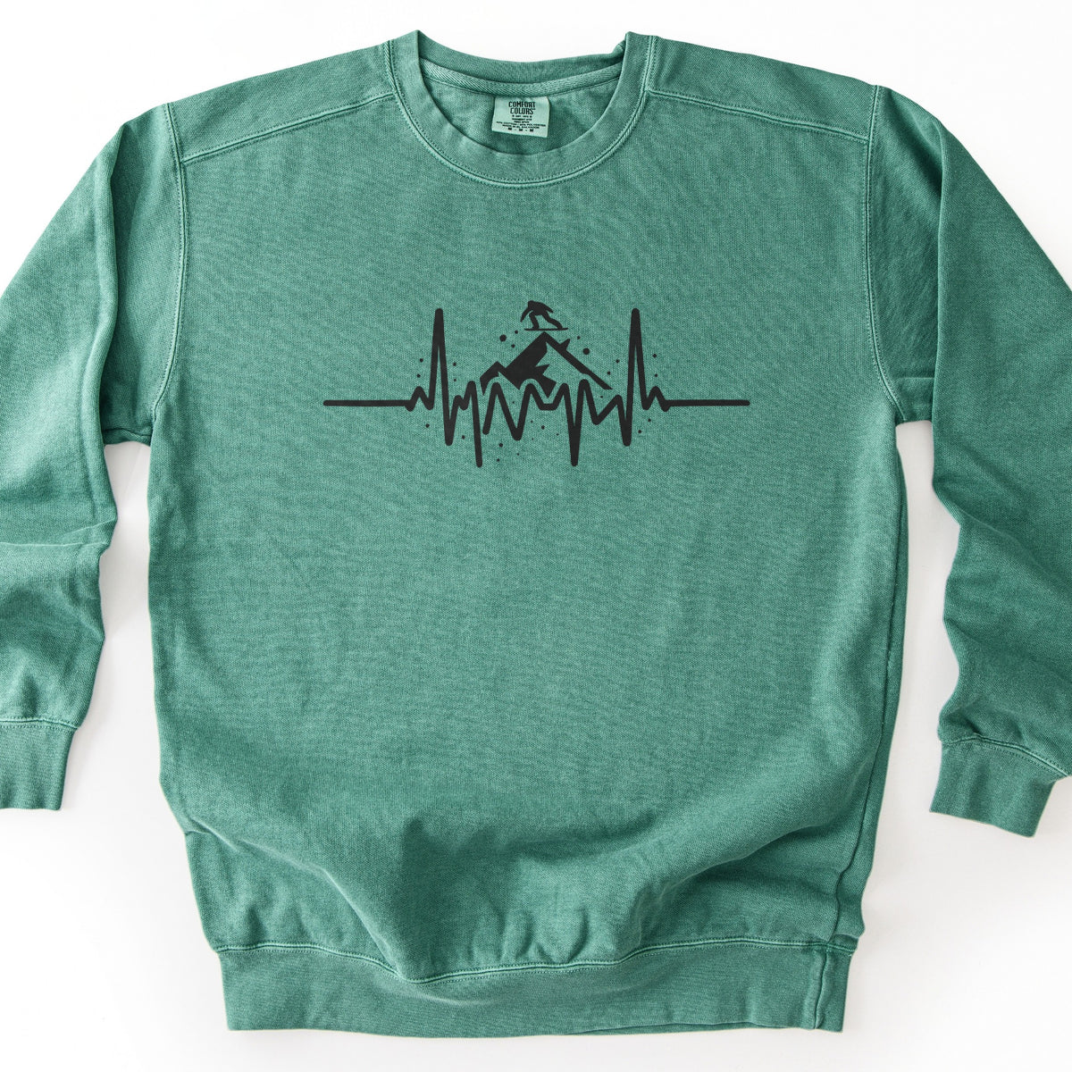 Snow Boarder Heartbeat Comfort Colors crewneck sweatshirt in Light Green, laid flat. A black heartbeat line runs across the chest, rising into a mountain peak with a snowboarder riding on top and small dots of snow around it. No words. The sweatshirt lies on a white surface.