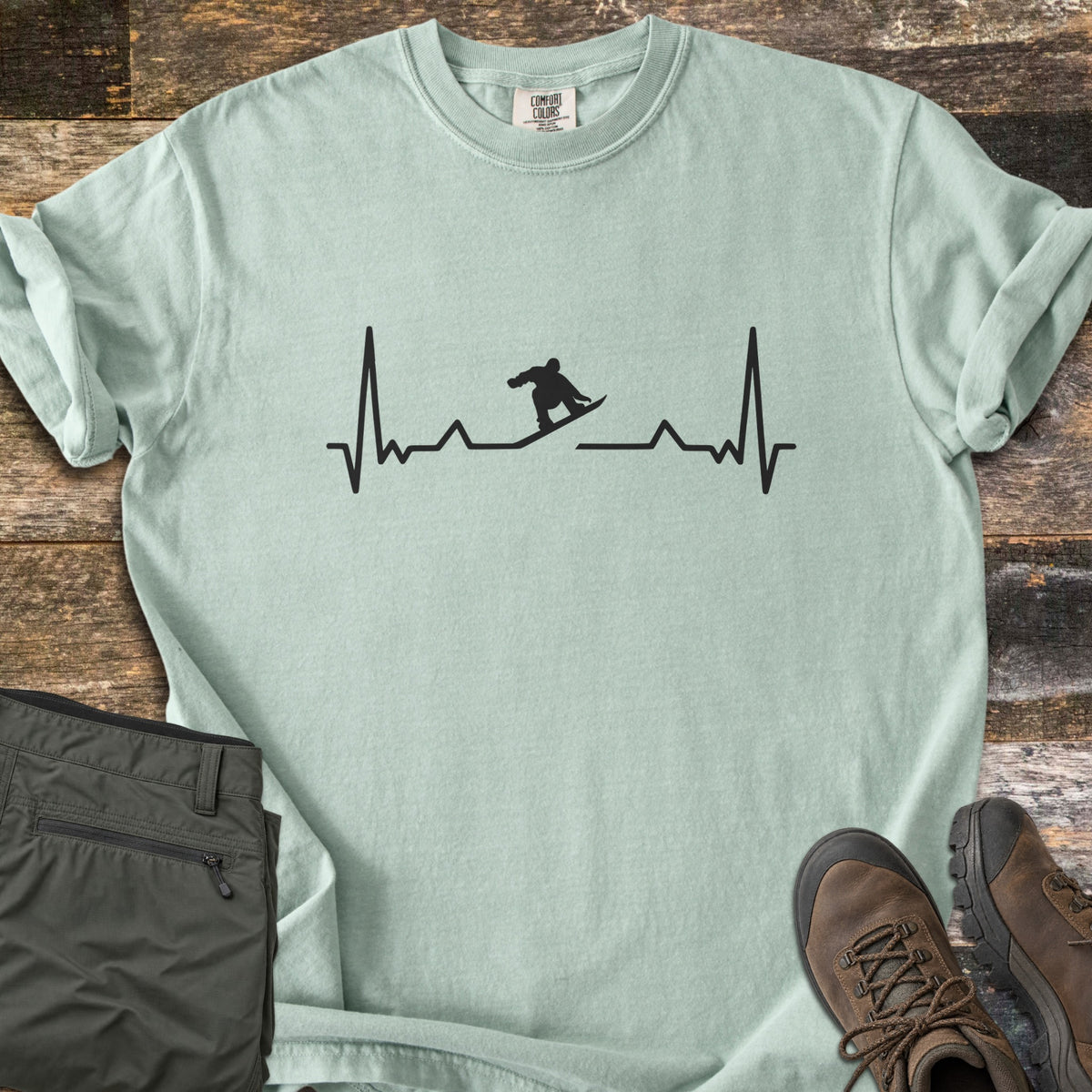 Snowboarder Heartbeat Comfort Colors T-shirt in Bay, laid flat. A black heartbeat line runs across the chest, with a snowboarder silhouette riding the line in the middle. Sleeves are rolled, on a rustic wood floor beside folded olive pants and brown hiking boots.