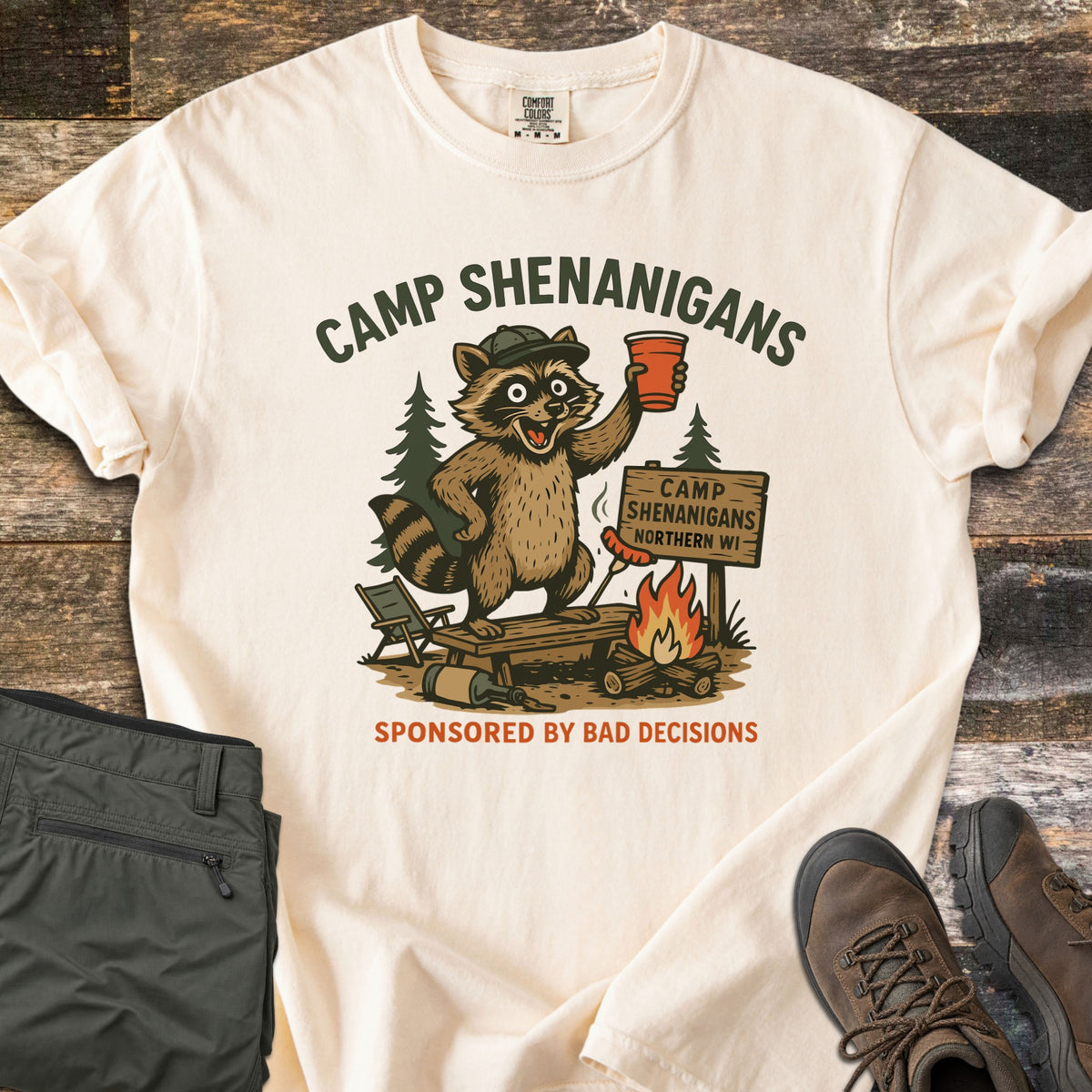 Camp Shenanigans Comfort Colors T-shirt in Ivory, laid flat. A raccoon in a cap raises a red cup by a campfire and a sign reading Camp Shenanigans Northern WI, under Camp Shenanigans and above Sponsored by Bad Decisions. Green pants and hiking boots sit on dark wood.