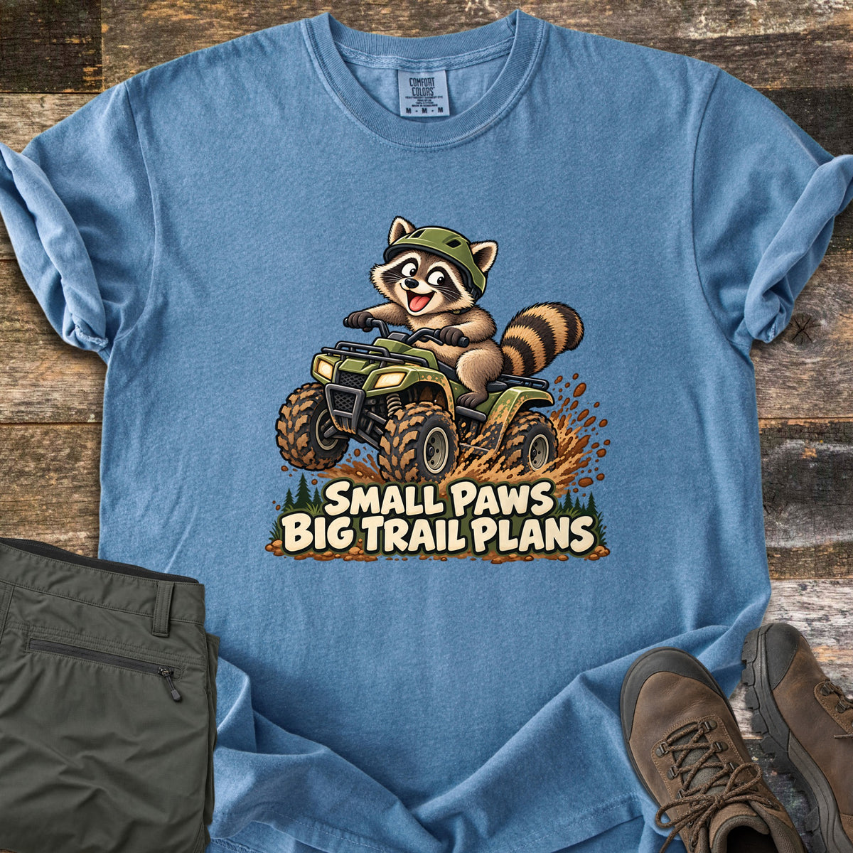 Small Paws Big Trail Plans Comfort Colors T-shirt in Blue Jean, laid flat. A cartoon raccoon in a green helmet rides a muddy green ATV, kicking up dirt above the words Small Paws Big Trail Plans. Shown on rustic wood with folded olive hiking pants and brown hiking boots.