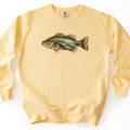 Rocky Shallows Bass Comfort Colors crewneck sweatshirt in Butter, laid flat. A large illustrated bass is printed across the chest, its side cut away to show clear shallow water with sunbeams over rounded rocks and pebbles. No words are printed. Shown on a plain white background.