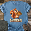 My Thanksgiving Contribution Comfort Colors T-shirt in Blue Jean, laid flat. A cartoon squirrel grins while holding a woven basket of acorns, a small pumpkin and autumn leaves above the words My Thanksgiving Contribution. Olive pants and brown hiking boots lie beside it on weathered wood.