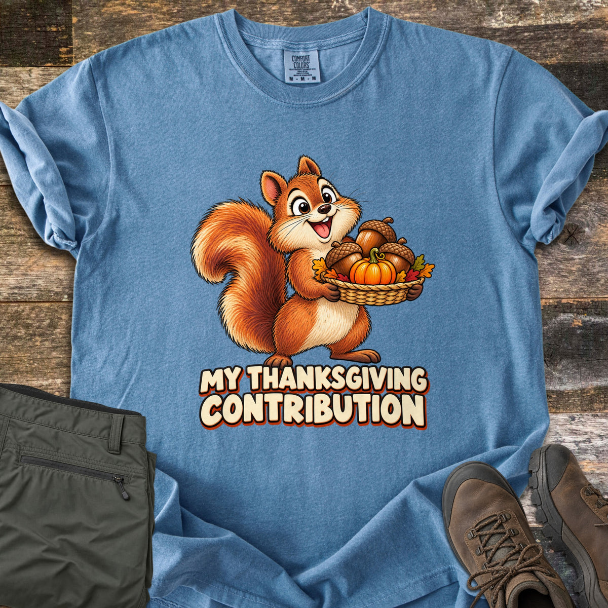 My Thanksgiving Contribution Comfort Colors T-shirt in Blue Jean, laid flat. A cartoon squirrel grins while holding a woven basket of acorns, a small pumpkin and autumn leaves above the words My Thanksgiving Contribution. Olive pants and brown hiking boots lie beside it on weathered wood.