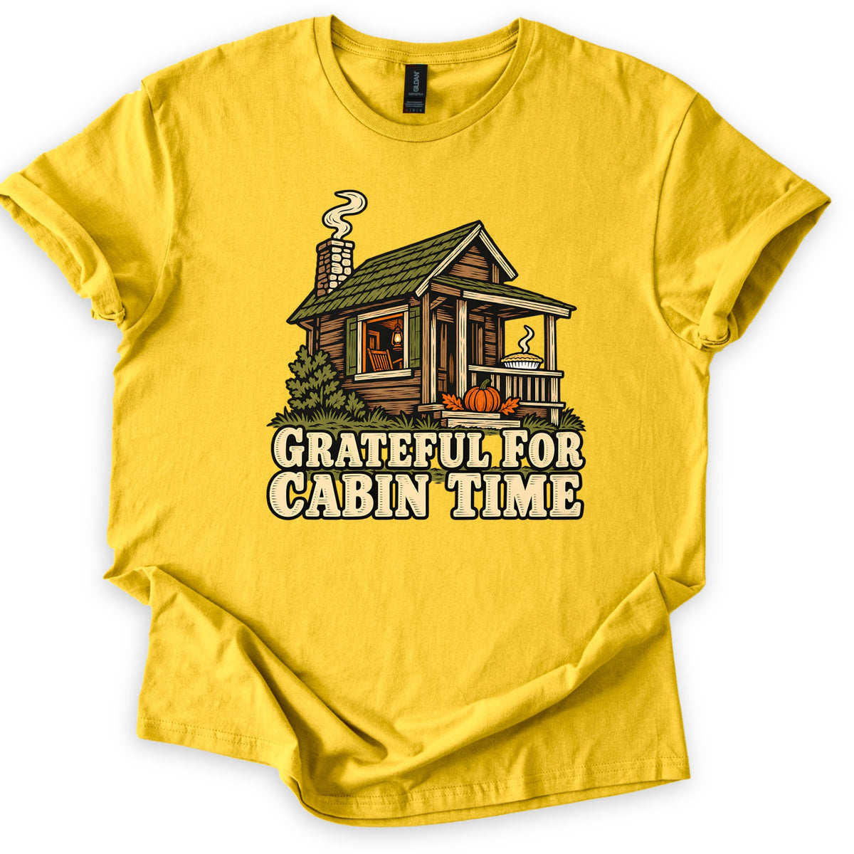 Grateful For Cabin Time Comfort Colors T-shirt in Daisy, laid flat. A log cabin with a smoking stone chimney, a lantern-lit window, a pie on the porch rail and a pumpkin on the steps sits above the words Grateful For Cabin Time. Sleeves rolled, on a white background.