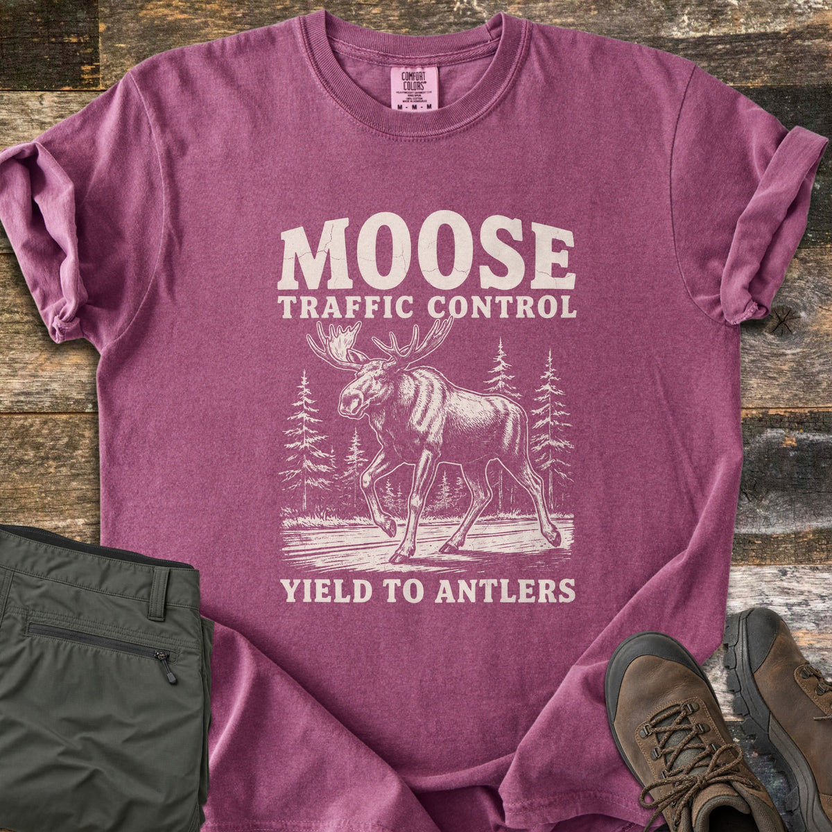 Moose Traffic Control Comfort Colors T-shirt in Berry, laid flat. An off-white sketch of a moose walking across a road between pine trees, with MOOSE TRAFFIC CONTROL above and YIELD TO ANTLERS below. Sleeves rolled, on weathered wood with olive pants and brown hiking boots.