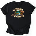 Watershed Engineer Comfort Colors T-shirt in Black, laid flat. A beaver whose body frames a scene of a snowy mountain, pine trees, a lake, and a log dam with a waterfall, between the words Watershed Engineer. The sleeves are rolled, on a white background.