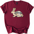 Snowshoe Hare Birch Woods Comfort Colors T-shirt in Maroon, laid flat. A painted snowshoe hare sits facing left, its body filled with a snowy birch forest, a winding path and distant mountains. No words are printed. The shirt lies on white with rolled sleeves.