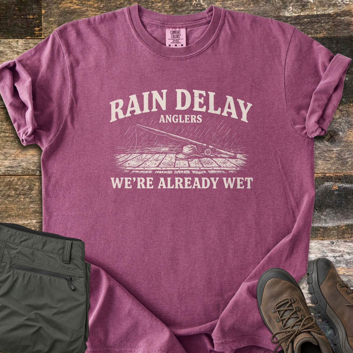Rain Delay Anglers Comfort Colors T-shirt in Berry, laid flat. Cream print of a fishing rod and bucket hat on a rain-soaked wooden dock, between the words Rain Delay Anglers and We're Already Wet. Sleeves rolled, on weathered boards with olive pants and brown hiking boots.