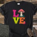 Mushroom Love Comfort Colors T-shirt in Black, laid flat. The word LOVE in distressed pink, lime green and orange letters with paint splatters, a red-capped mushroom on grass as the O. Shown on rustic wood with olive cargo pants and brown hiking boots.