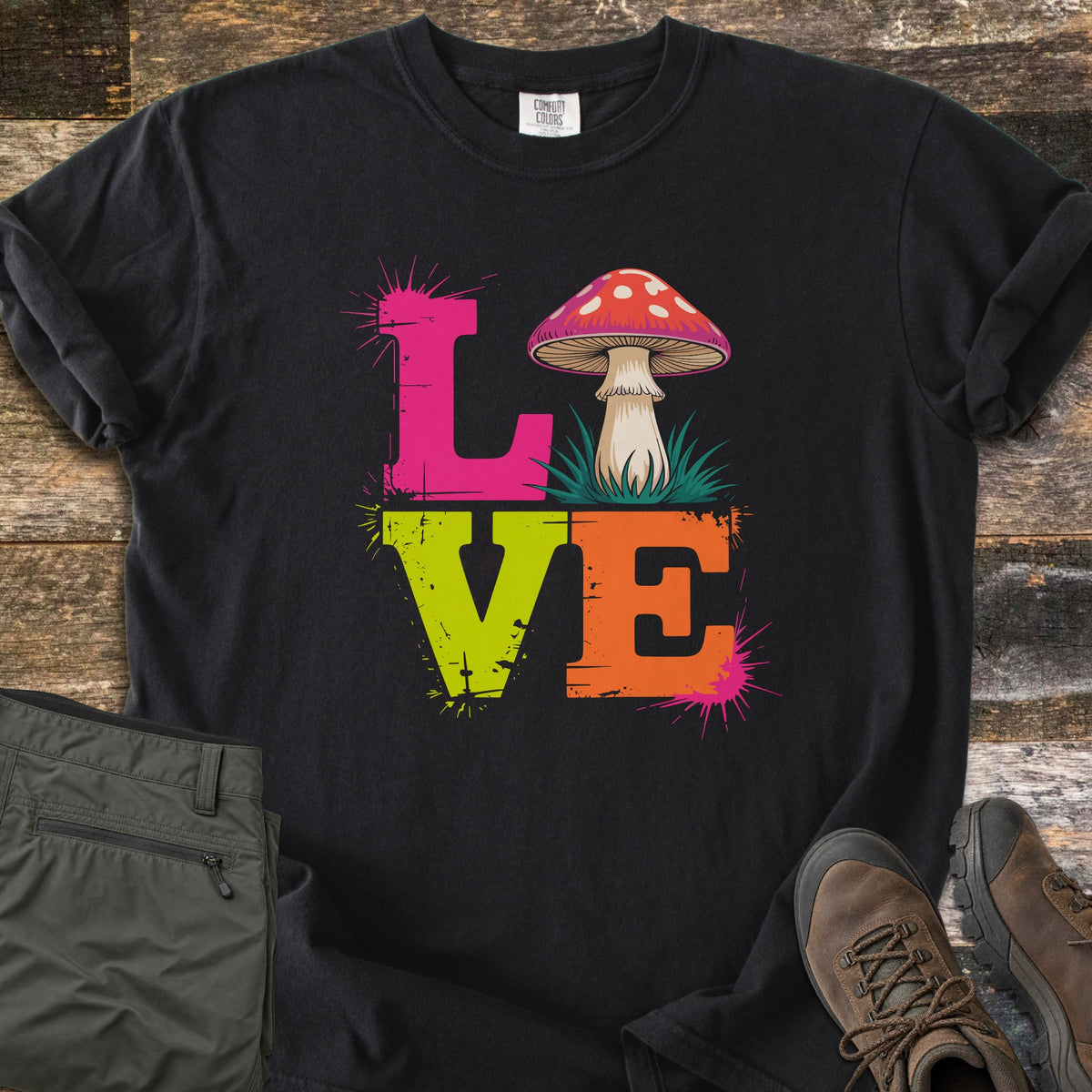 Mushroom Love Comfort Colors T-shirt in Black, laid flat. The word LOVE in distressed pink, lime green and orange letters with paint splatters, a red-capped mushroom on grass as the O. Shown on rustic wood with olive cargo pants and brown hiking boots.