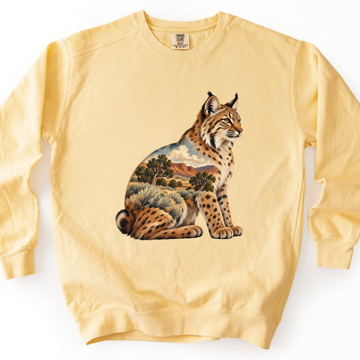Bobcat Sagebrush Canyon Comfort Colors crewneck sweatshirt in Butter, laid flat. A seated bobcat with spotted fur and tufted ears looks to the right; its body is filled with a painted desert scene of sagebrush, juniper trees, red canyon cliffs and a cloudy blue sky. No words are printed.