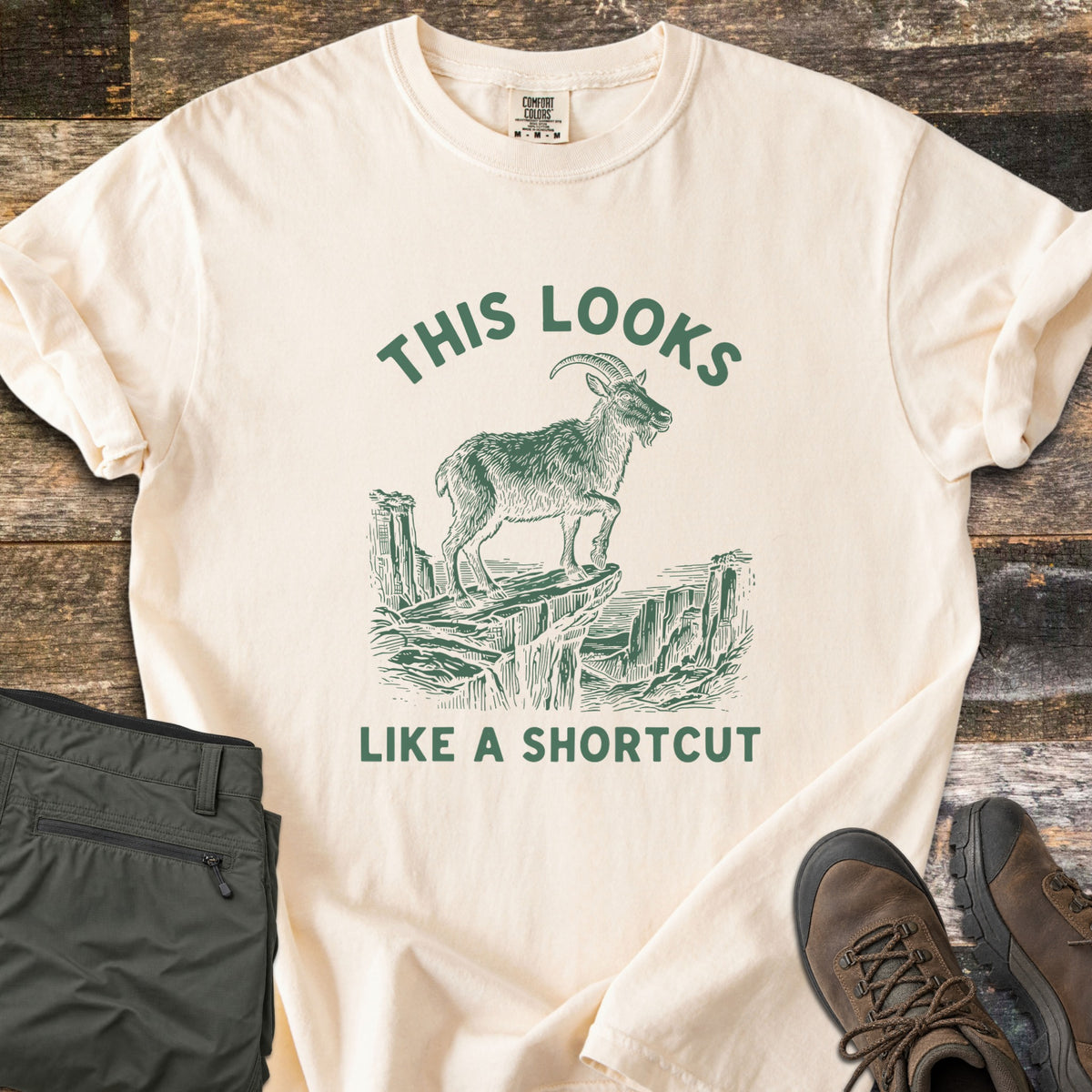 This Looks Like A Shortcut Comfort Colors T-shirt in Ivory, laid flat. A dark green etching of a horned goat standing on a cliff ledge over a canyon, with the words This Looks arched above and Like A Shortcut below. Olive hiking pants and brown hiking boots lie beside it on dark wood.