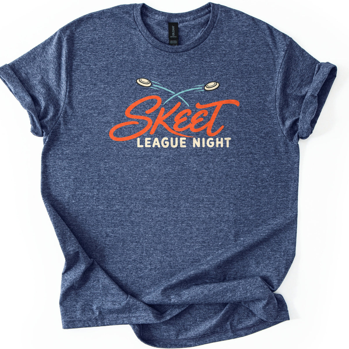 Skeet League Night Comfort Colors T-shirt in Heather Navy, laid flat. Two cream clay targets with crossing teal trails fly above the word Skeet in orange script, with League Night in cream capitals below. The shirt has rolled sleeves on a white background.