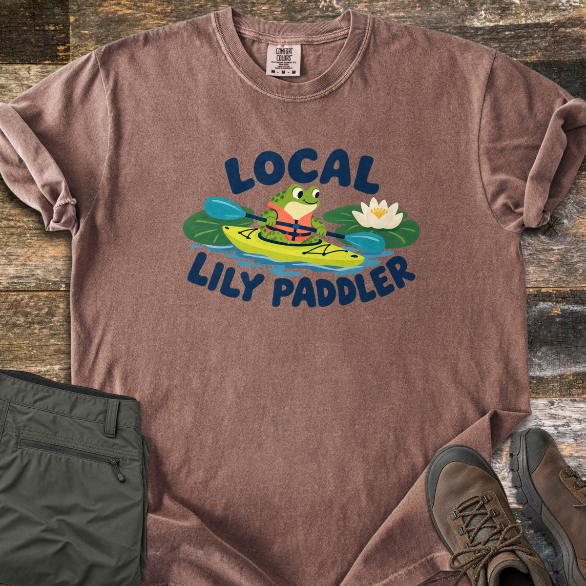 Lily Paddler Comfort Colors T-shirt in Espresso, laid flat. A cartoon frog in an orange life vest paddles a yellow kayak between lily pads and a white water lily, with the words Local Lily Paddler. Folded olive hiking pants and brown hiking boots sit on a wood plank background.