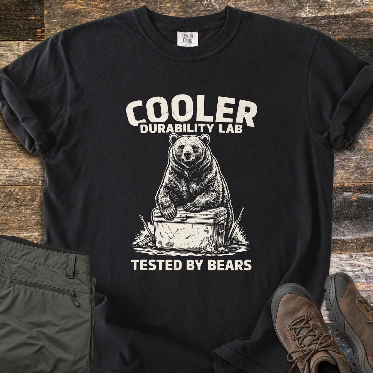 Cooler Durability Lab Comfort Colors T-shirt in Black, laid flat. A bear rests its paws on a cracked cooler between the words Cooler Durability Lab and Tested By Bears, in white. Olive hiking pants and brown boots sit beside it on weathered wood boards.