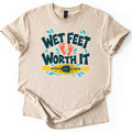 Wet Feet Worth It Comfort Colors T-shirt in Sand, laid flat. Dark teal bubbly letters read Wet Feet Worth It, with two orange footprints between the lines and a yellow kayak and paddle below, ringed by blue water splashes. The shirt lies on a white background.