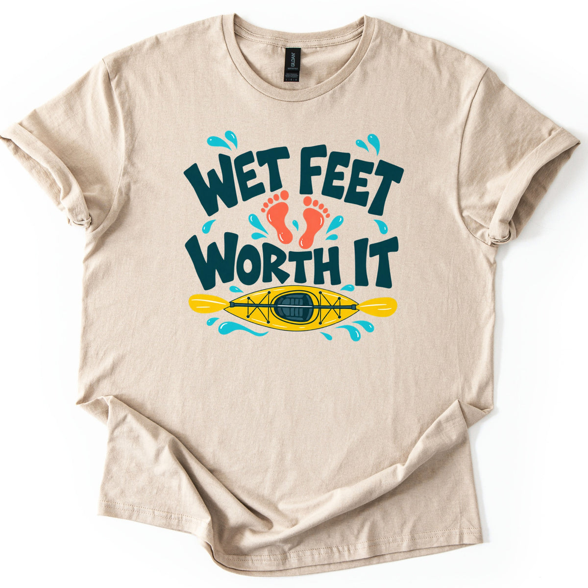 Wet Feet Worth It Comfort Colors T-shirt in Sand, laid flat. Dark teal bubbly letters read Wet Feet Worth It, with two orange footprints between the lines and a yellow kayak and paddle below, ringed by blue water splashes. The shirt lies on a white background.