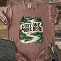 One More Bend Comfort Colors T-shirt in Espresso, laid flat. A woodcut-style scene of a winding river through pine forest and mountains, with a yellow canoe, and the words Just One More Bend. Sleeves rolled, on weathered wood beside olive hiking pants and brown boots.