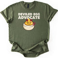 Deviled Egg Advocate Comfort Colors T-shirt in Military Green, laid flat. Bold cream text reads Deviled Egg Advocate above a cartoon deviled egg with red devil horns, a wink, and a small red pitchfork. The shirt has cuffed sleeves and lies on a white background.