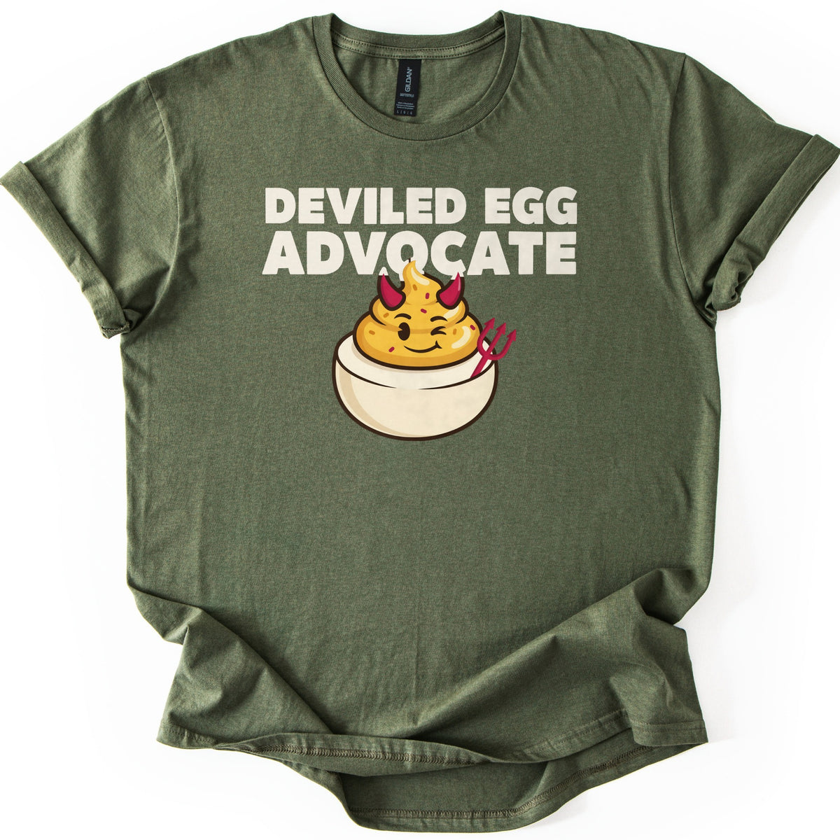 Deviled Egg Advocate Comfort Colors T-shirt in Military Green, laid flat. Bold cream text reads Deviled Egg Advocate above a cartoon deviled egg with red devil horns, a wink, and a small red pitchfork. The shirt has cuffed sleeves and lies on a white background.