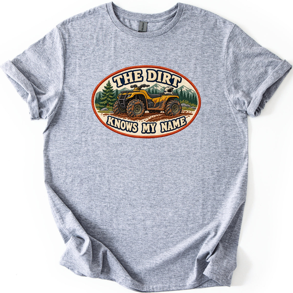 The Dirt Knows My Name Comfort Colors T-shirt in Sport Grey, laid flat. An oval patch-style print shows a mud-covered yellow ATV with a white helmet, pine trees and mountains, with THE DIRT above and KNOWS MY NAME below. The shirt lies on a white background with sleeves cuffed.