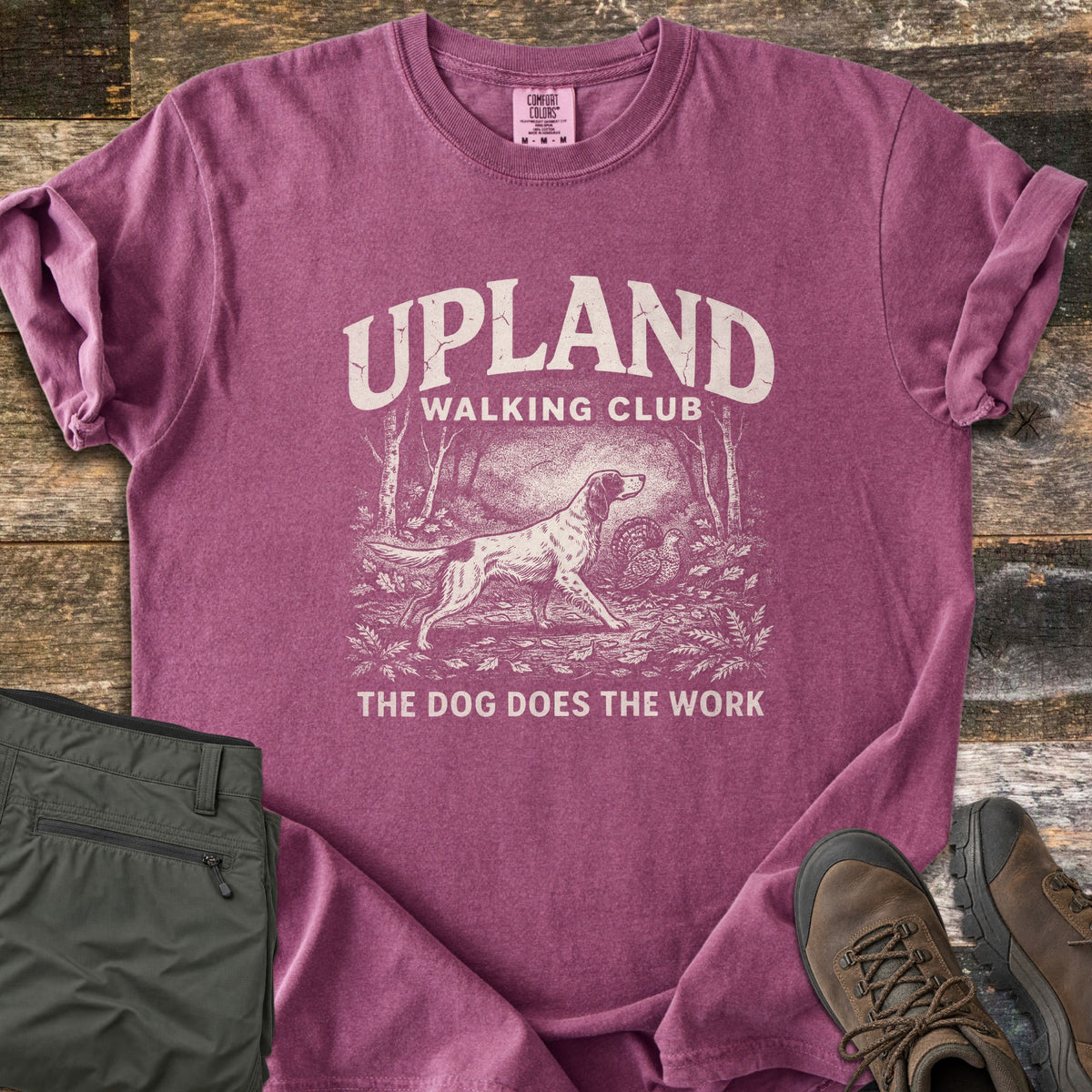 Upland Walking Club Comfort Colors T-shirt in Berry, laid flat. A vintage-style sketch of a spaniel pointing at a grouse in the woods, under the words Upland Walking Club and above The Dog Does The Work. Olive pants and brown hiking boots sit beside it on weathered wood.