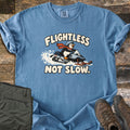 Flightless Not Slow Comfort Colors T-shirt in Blue Jean, laid flat. A cartoon penguin in a helmet and red scarf rides a snowmobile through snow between the words Flightless Not Slow. Folded olive pants and brown hiking boots sit beside it on weathered wood.