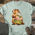 Groundhog Blossom Meadow Comfort Colors T-shirt in Bay, laid flat. A painted groundhog sits upright, its body filled with a spring meadow scene of pink blossoming trees, green hills and blue sky; no words. Sleeves rolled, on weathered wood beside folded olive pants and brown hiking boots.