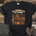 Drunked Up II Comfort Colors T-shirt in Black, laid flat. A cartoon bear in a cap lounges in a camp chair by a campfire, raising a red cup, under the words All Drunked Up at Camp, with a sign reading Camp Nowhere Up. Olive pants and hiking boots lie on weathered wood.