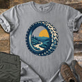 Knobby Tire Trail Comfort Colors T-shirt in Grey, laid flat. A mountain bike tire frames a scene of a winding trail through green hills and pine trees toward mountains and a yellow sun. Olive cargo pants and brown hiking boots lie beside it on weathered wood planks.