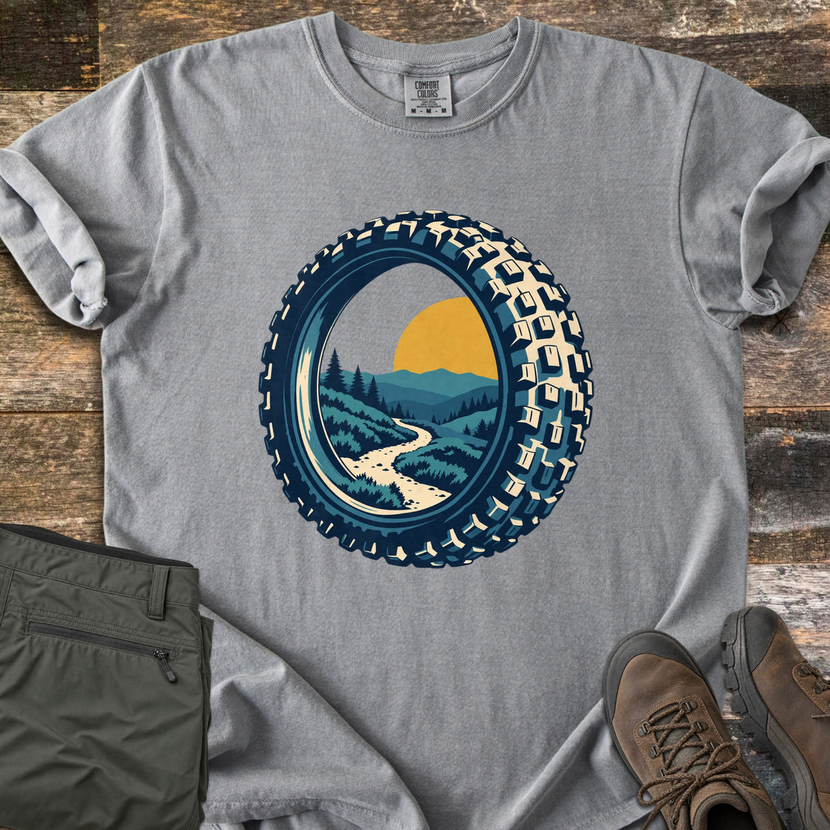 Knobby Tire Trail Comfort Colors T-shirt in Grey, laid flat. A mountain bike tire frames a scene of a winding trail through green hills and pine trees toward mountains and a yellow sun. Olive cargo pants and brown hiking boots lie beside it on weathered wood planks.