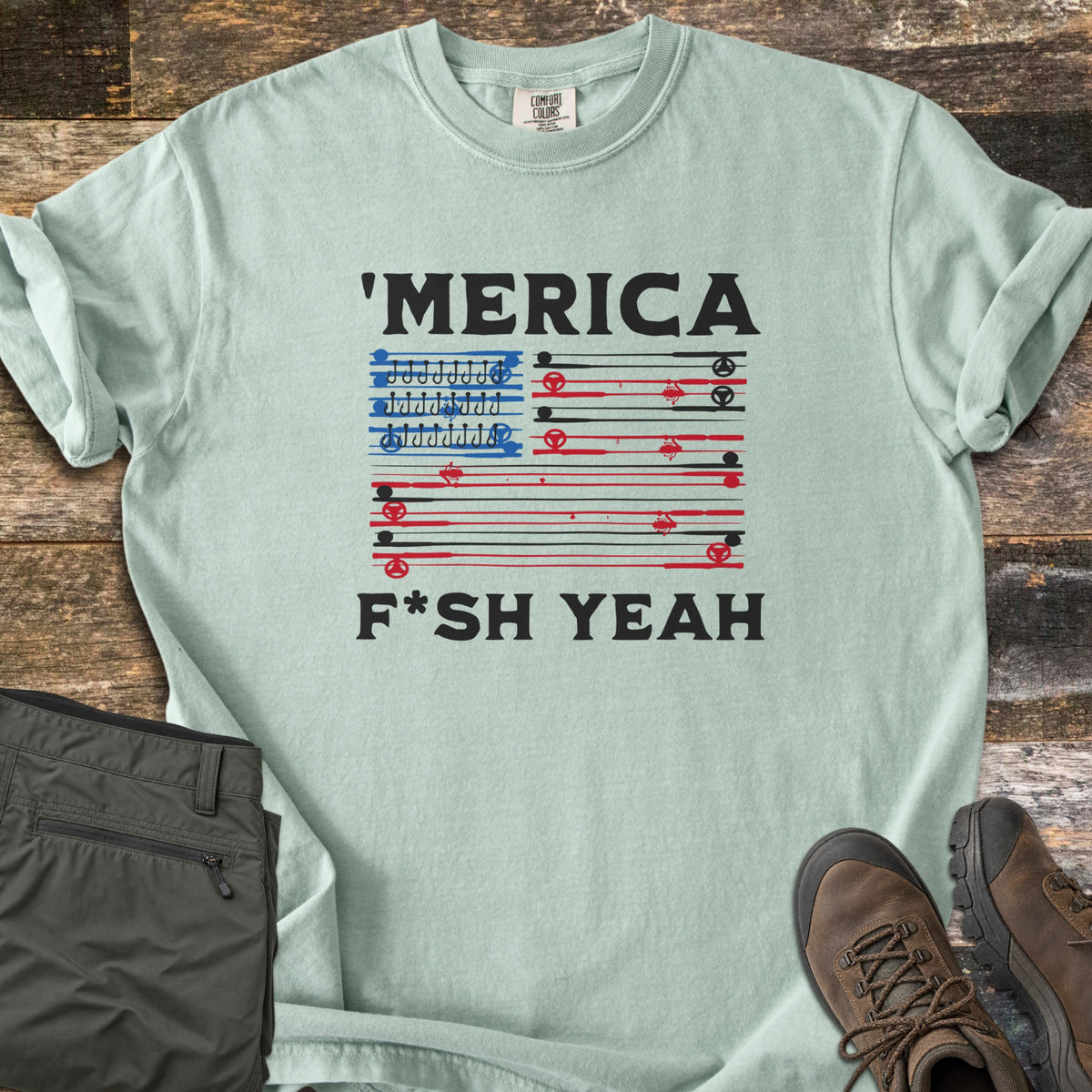 Fish America Fish Yeah Comfort Colors T-shirt in Bay, laid flat. Black text reads 'MERICA above an American flag made of red and black fishing rods, with rows of hooks in the blue corner, then F*SH YEAH. Folded olive pants and brown hiking boots sit on weathered wood.