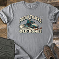 Fresh Tracks Old Bones Comfort Colors T-shirt in Grey, laid flat. A skeleton in a helmet and goggles rides a teal and cream snowmobile through spraying snow, between the words Fresh Tracks and Old Bones. Olive hiking pants and brown boots sit beside it on weathered wood.