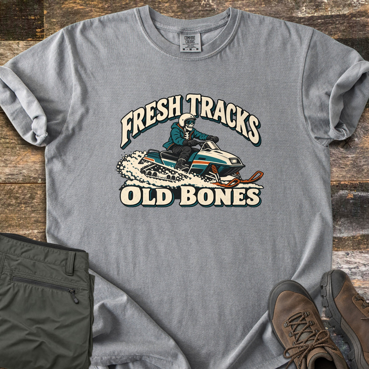 Fresh Tracks Old Bones Comfort Colors T-shirt in Grey, laid flat. A skeleton in a helmet and goggles rides a teal and cream snowmobile through spraying snow, between the words Fresh Tracks and Old Bones. Olive hiking pants and brown boots sit beside it on weathered wood.