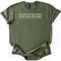 I Know Every Bird in My Yard by Name Comfort Colors T-shirt in Military Green, laid flat. Cream serif text across the chest reads I KNOW EVERY BIRD IN MY YARD BY NAME. THEY DON'T KNOW MINE. The sleeves are cuffed and the shirt lies on a plain white background.